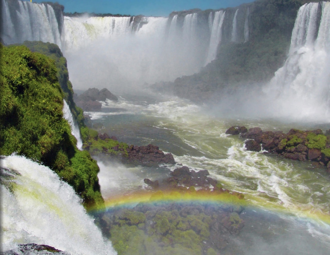 Foz do Iguaçu: vista aproximada das Cataratas do Iguaçu, com múltiplas quedas sobre paredões rochosos cobertos de mata e um arco-íris na névoa