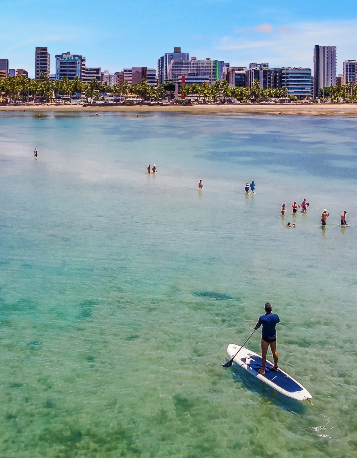 Maceió: vista aérea de praia urbana de águas claras e rasas, com coqueiros, prédios da orla e pessoa em pé num stand-up paddle