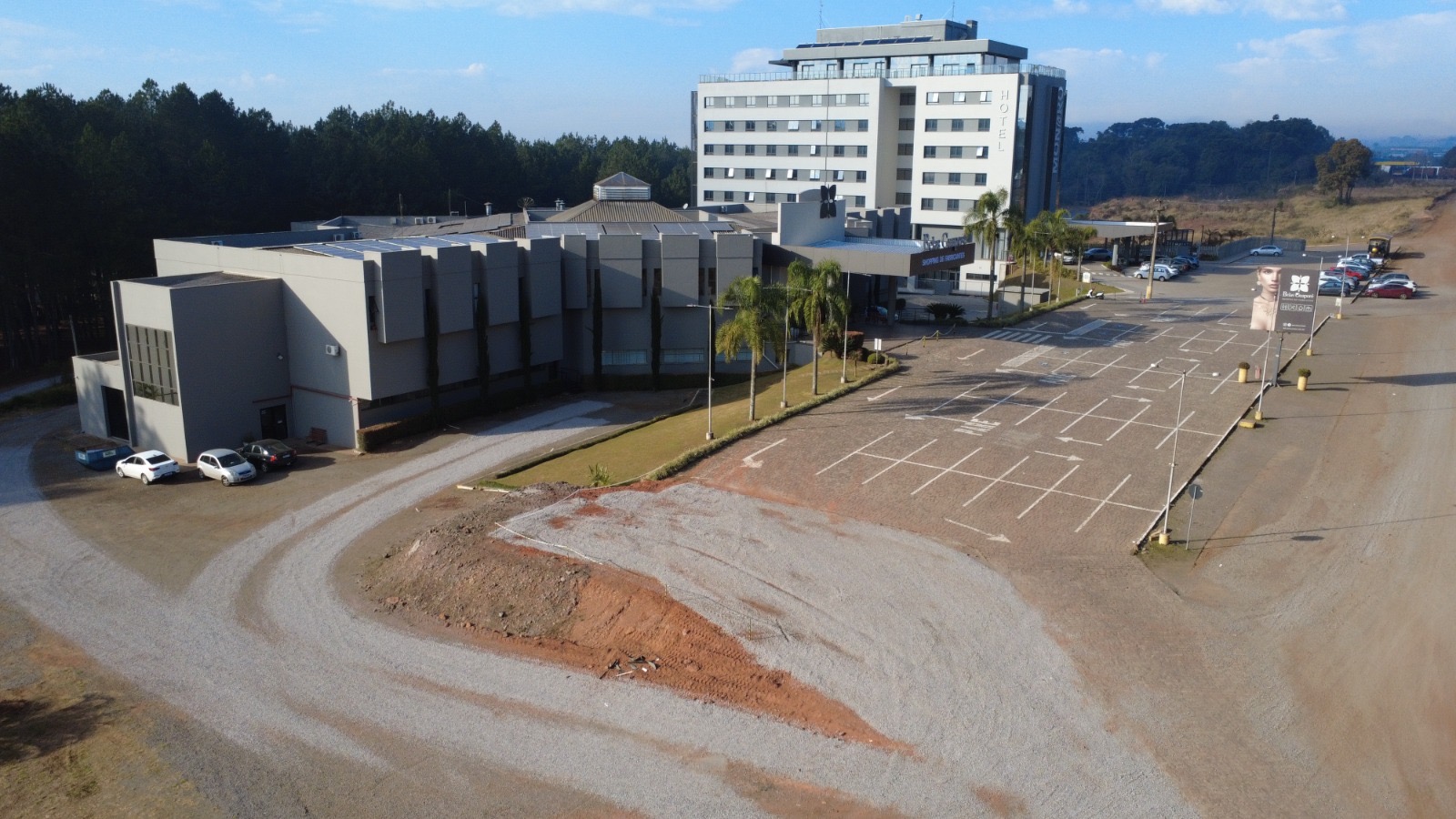 Vista aérea por drone de área em preparo com estrada de terra, estacionamento vazio e o prédio do Belas Guaporé Shopping de Fabricantes ao fundo