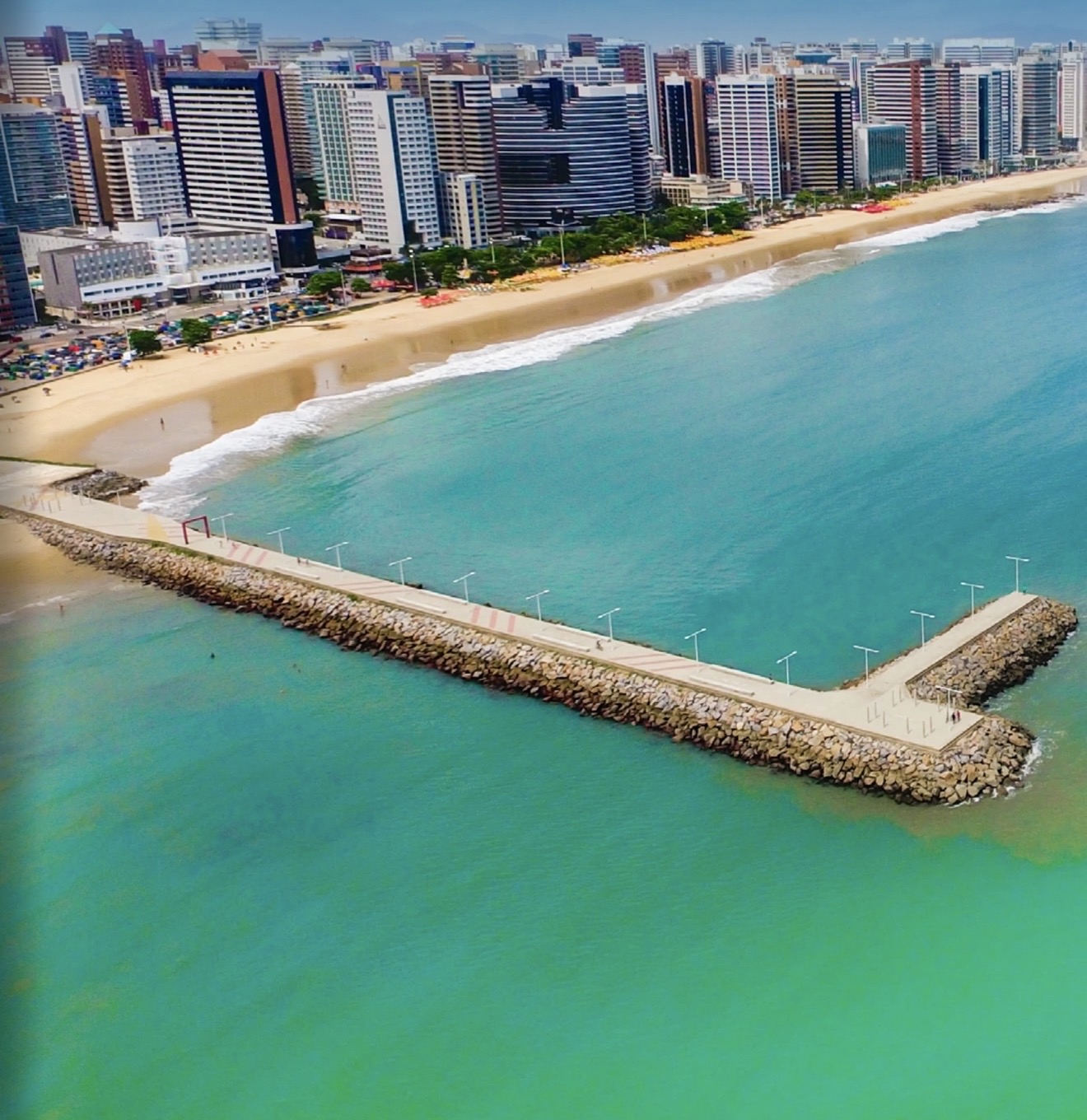 Fortaleza: vista aérea do espigão de pedras com passarela sobre o mar verde-turquesa, com a orla urbana de prédios ao fundo