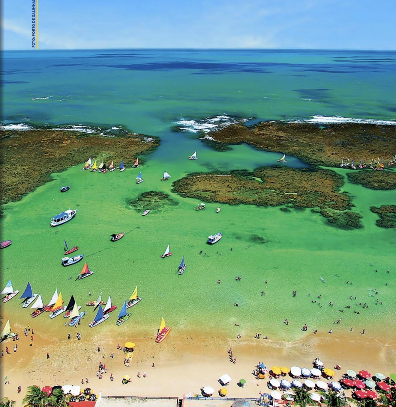 Porto de Galinhas: vista aérea das piscinas naturais entre arrecifes, com jangadas de vela coloridas, barcos, banhistas e guarda-sóis na praia